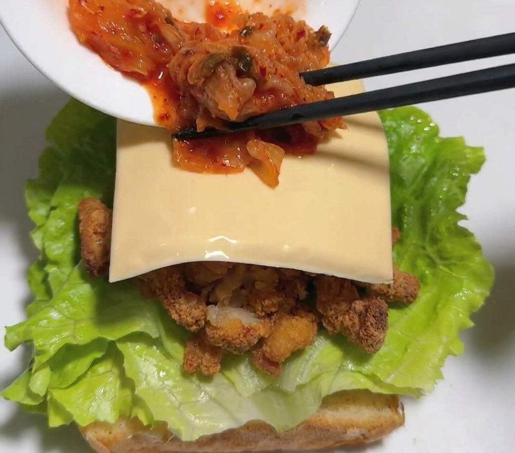 Palillos negros colocando kimchi de col roja sobre una loncha de queso dentro del sándwich.