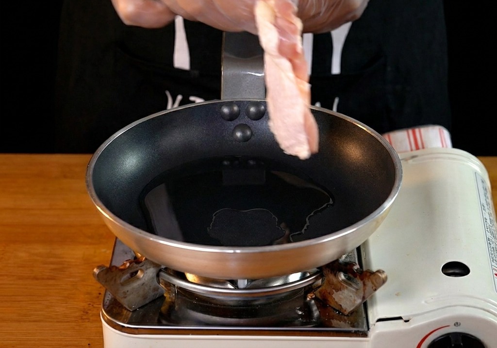 Una mano colocando un muslo de pollo crudo en una sartén con aceite caliente.