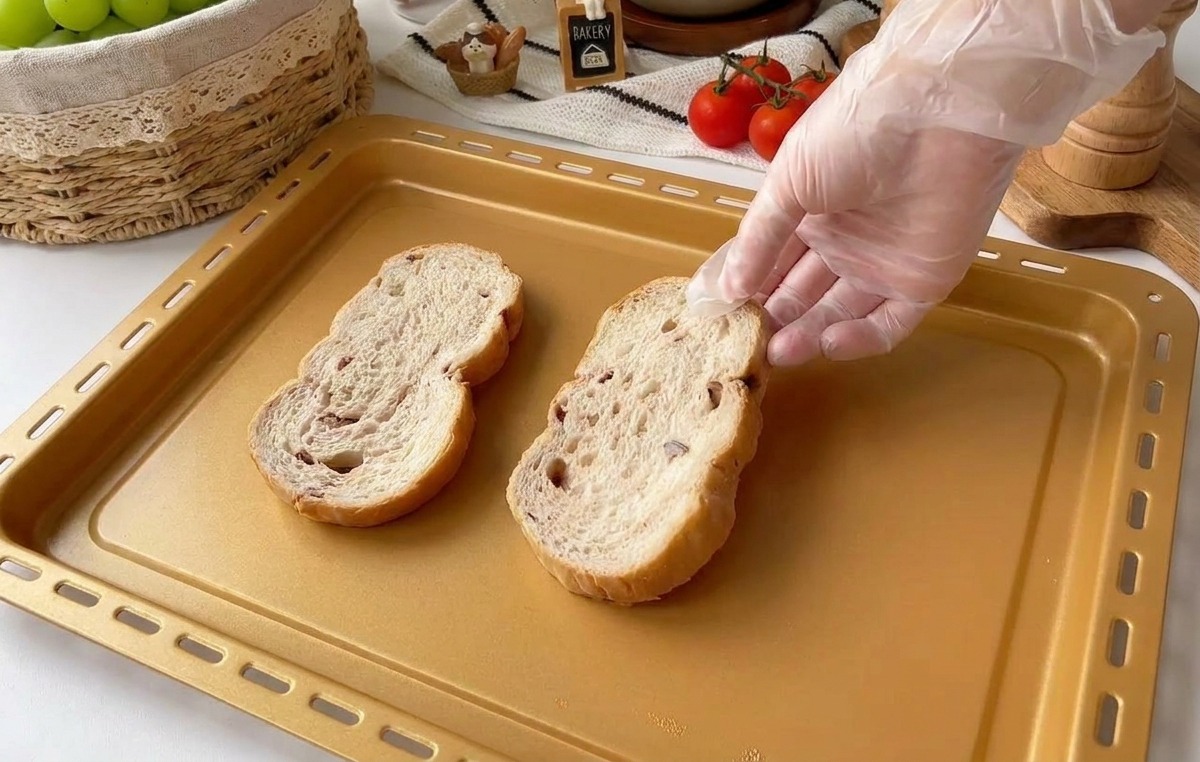 Una mano con guante coloca dos rebanadas gruesas de pan rústico multigrano en una bandeja de horno dorada.