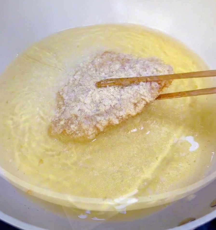 Un filete de pollo empanizado sumergiéndose en una sartén con aceite caliente brillante usando palillos de madera.