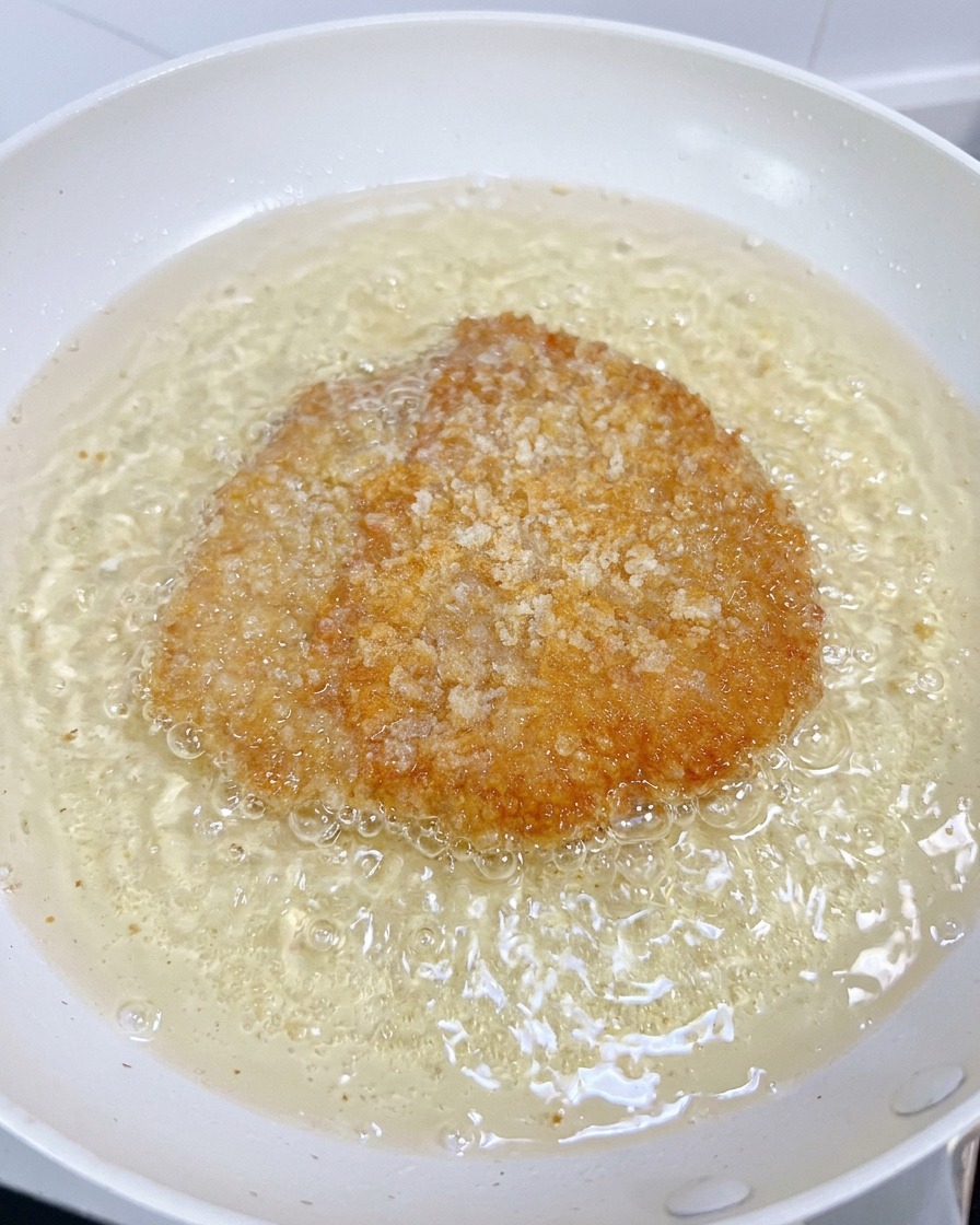 Un filete de pollo friéndose en una sartén blanca con aceite burbujeante, adquiriendo un color dorado y crujiente.
