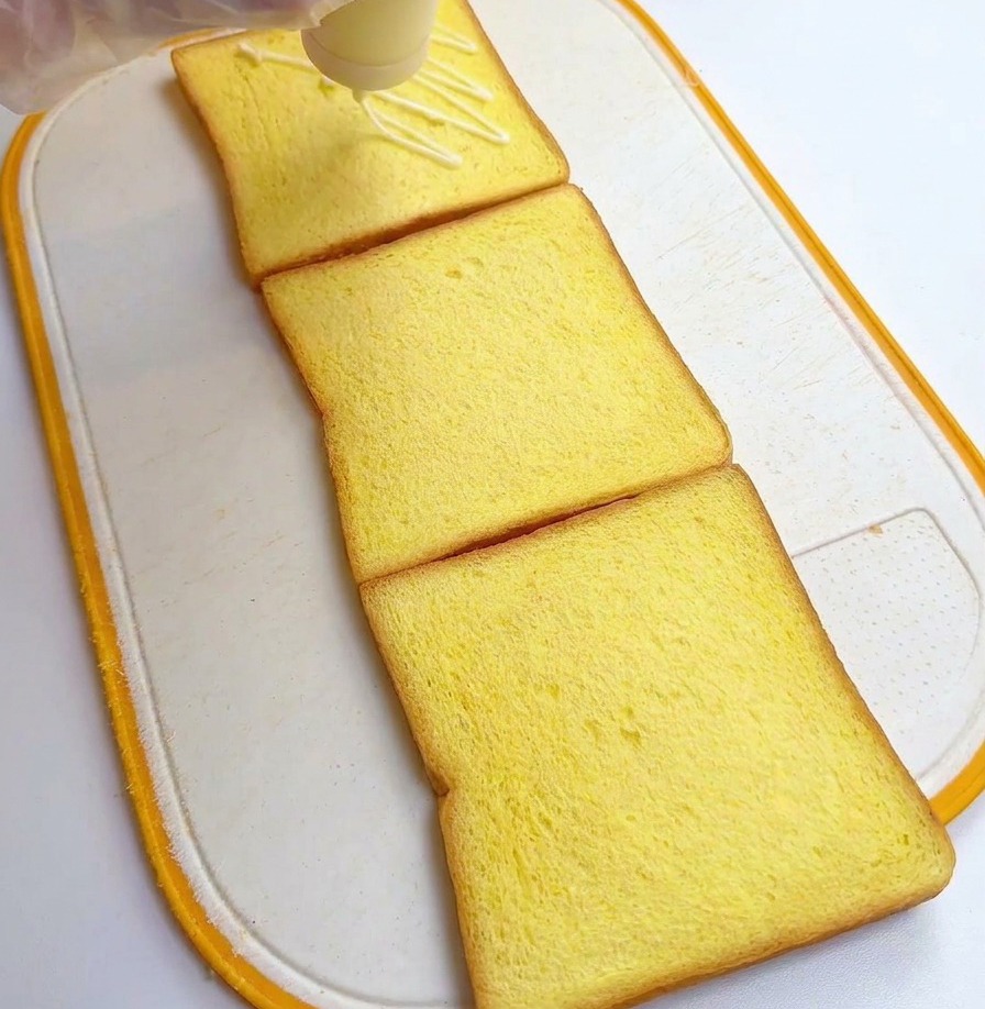 Una persona con guantes de plástico rociando aderezo blanco desde una botella dispensadora sobre tres rebanadas de pan de molde amarillo.