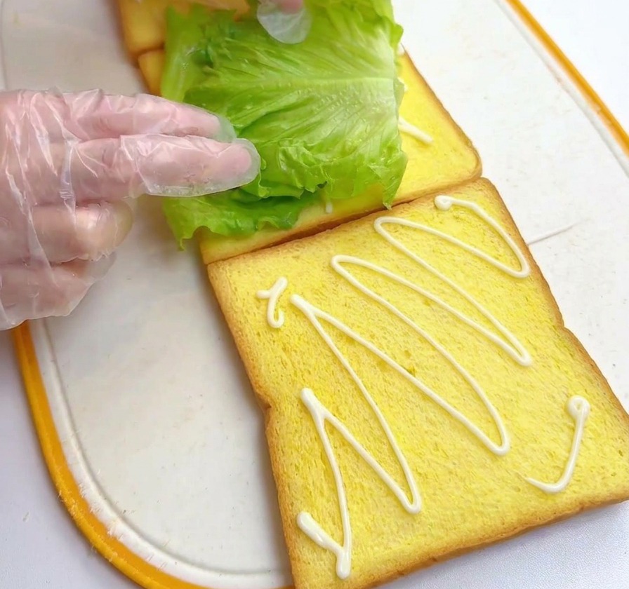 Manos colocando una hoja de lechuga verde y vibrante sobre una rebanada de pan amarillo previamente aderezada.