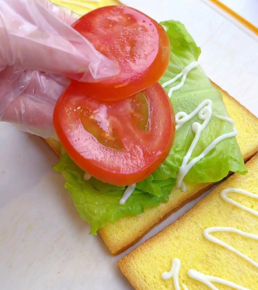 Una persona con guantes de plástico colocando dos rodajas redondas de tomate sobre una cama de lechuga verde en una rebanada de pan.