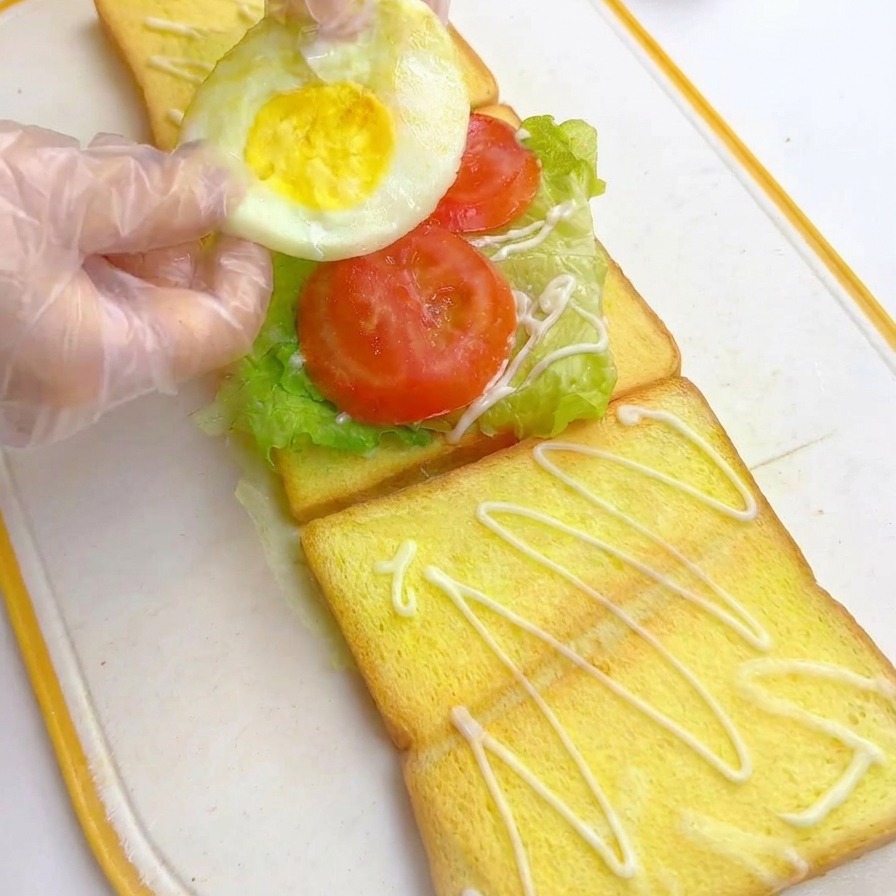 Manos enguantadas colocando un huevo frito redondo sobre las rodajas de tomate en el montaje de un sándwich de varias capas.