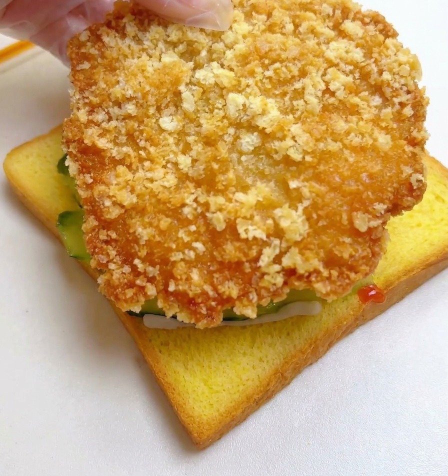 Una mano colocando un filete de pollo grande, empanizado y dorado sobre la rebanada de pan con pepino.