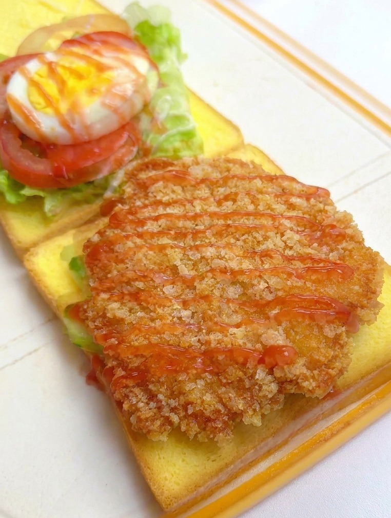 Un filete de pollo empanizado y dorado sobre un pan siendo rociado con kétchup rojo, con otras capas del sándwich visibles al fondo.