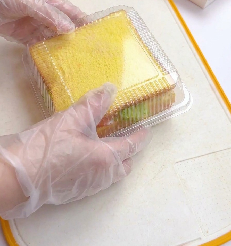 Manos con guantes de plástico presionando la tapa cuadrada de plástico transparente sobre un sándwich grueso apilado dentro de un molde.