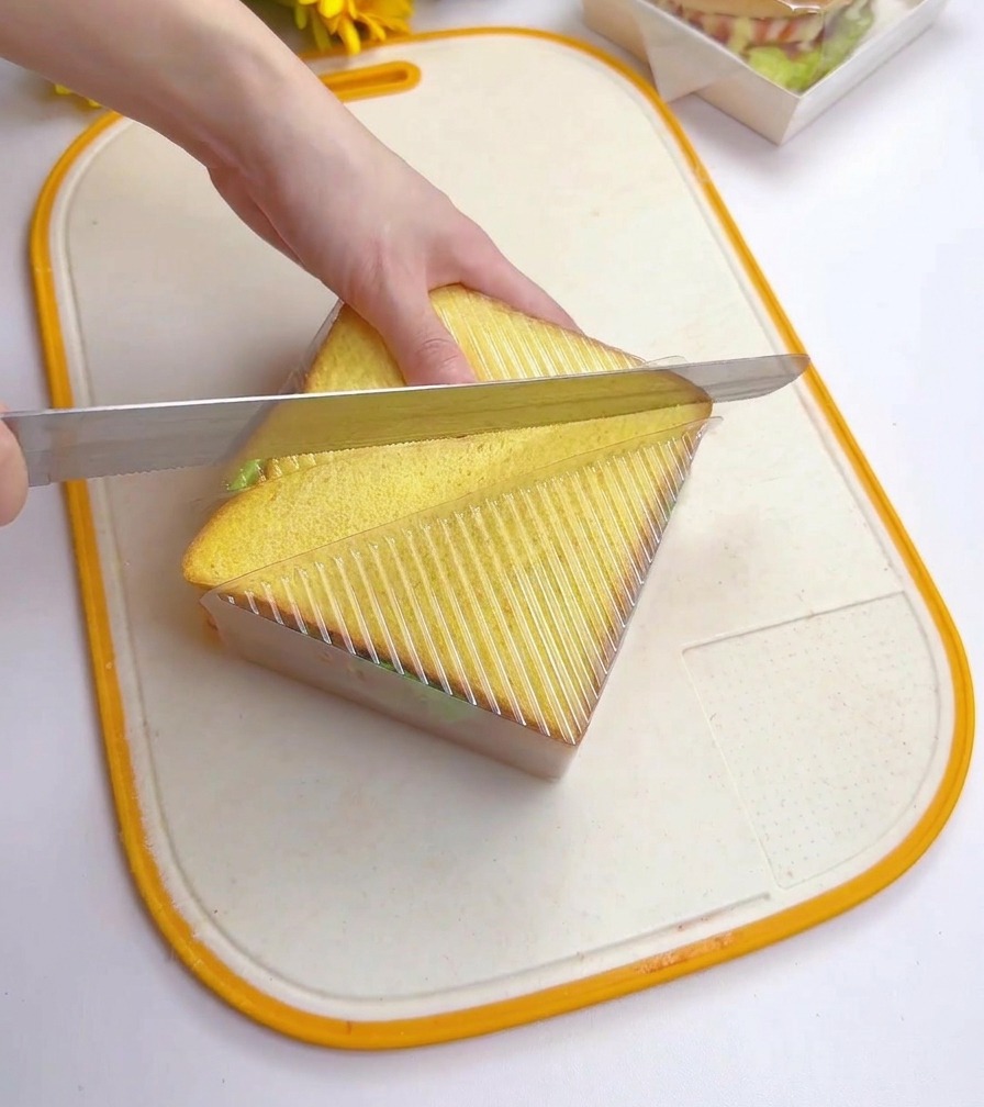 Una persona usando un cuchillo de sierra para cortar un sándwich apilado en diagonal mientras está sujeto dentro de un recipiente de plástico triangular transparente.