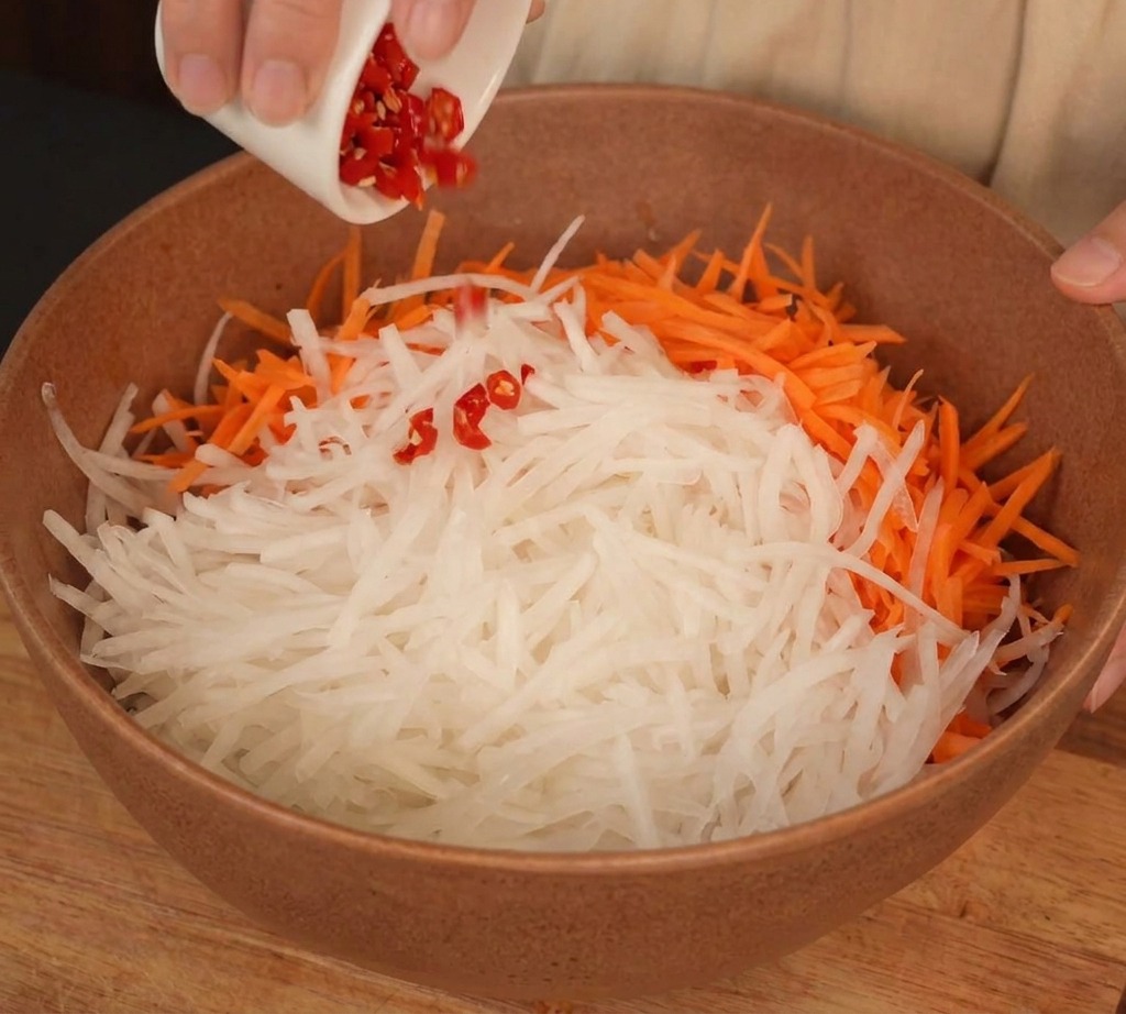 Un bol que contiene rábano blanco rallado y zanahorias naranjas a las que se les añaden chiles rojos picados desde un pequeño plato blanco.