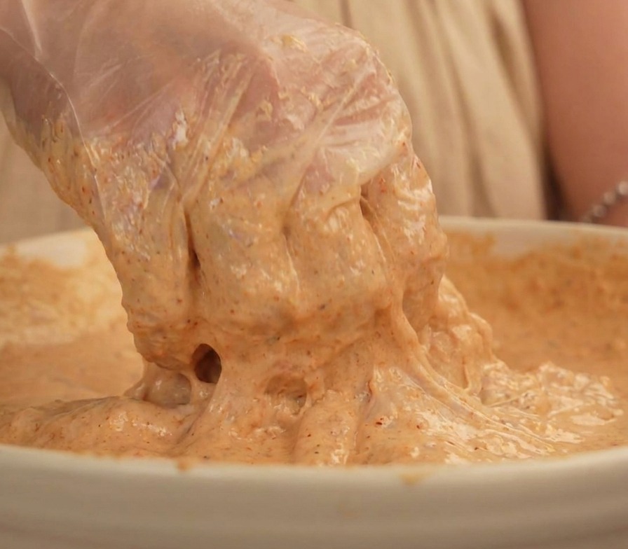 Una mano con un guante de plástico transparente levantando un trozo de muslo de pollo completamente cubierto por un rebozado espeso de color beige.
