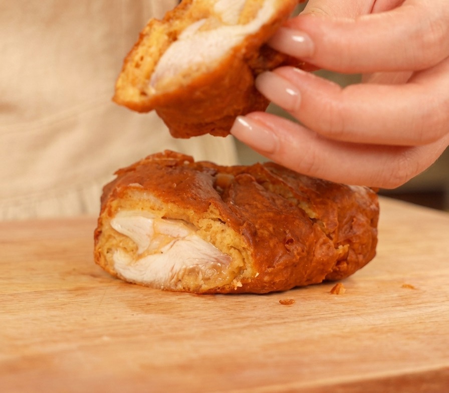 Un trozo de pollo frito cortado en una tabla de cortar de madera, mostrando la carne blanca y jugosa y una costra dorada, gruesa y crujiente.