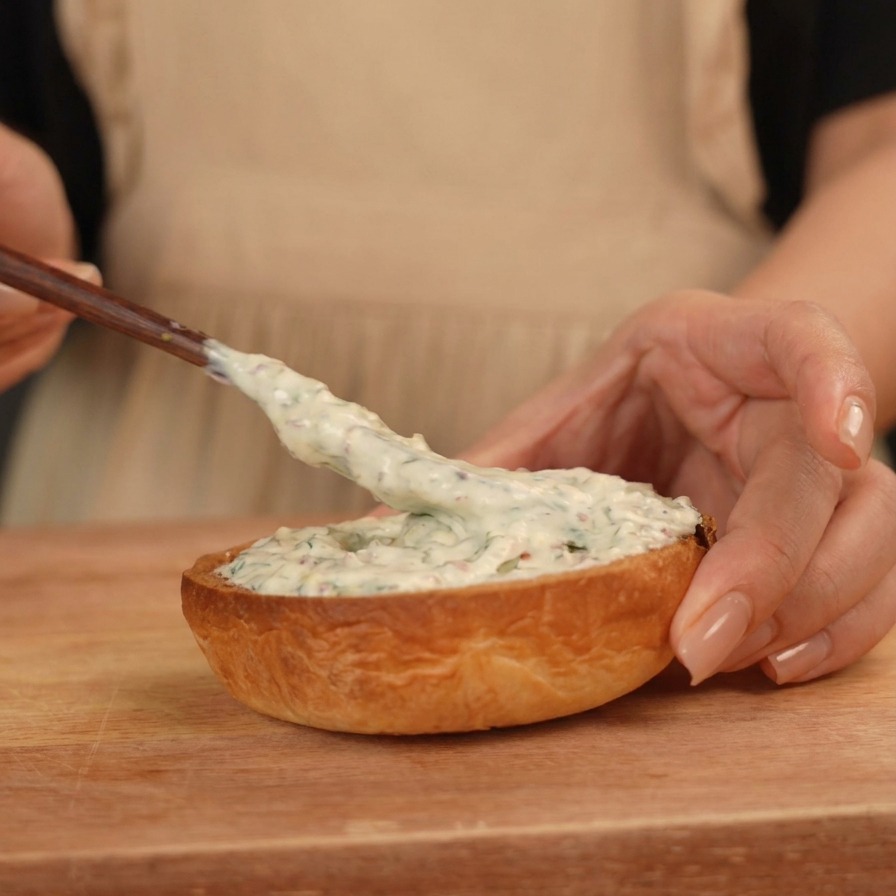 Una mano usando un pequeño esparcidor de madera para aplicar una salsa blanca, espesa y cremosa, en la mitad inferior de un pan de hamburguesa tostado.