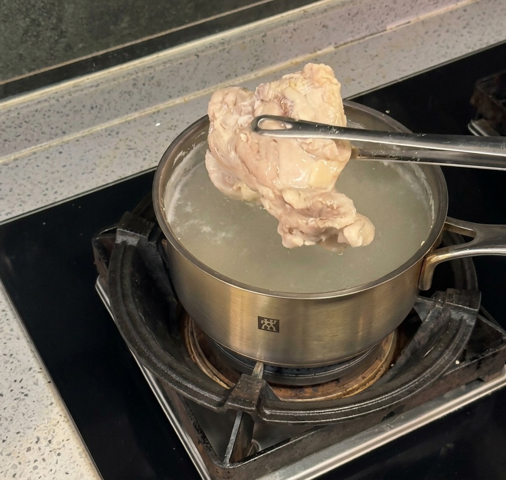 Unas pinzas de metal levantando una pieza grande de pollo cocido de una olla de acero inoxidable con agua hirviendo.