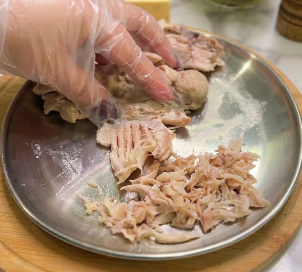 Una mano con guante desmenuzando pollo cocido en tiras finas sobre un plato de metal.