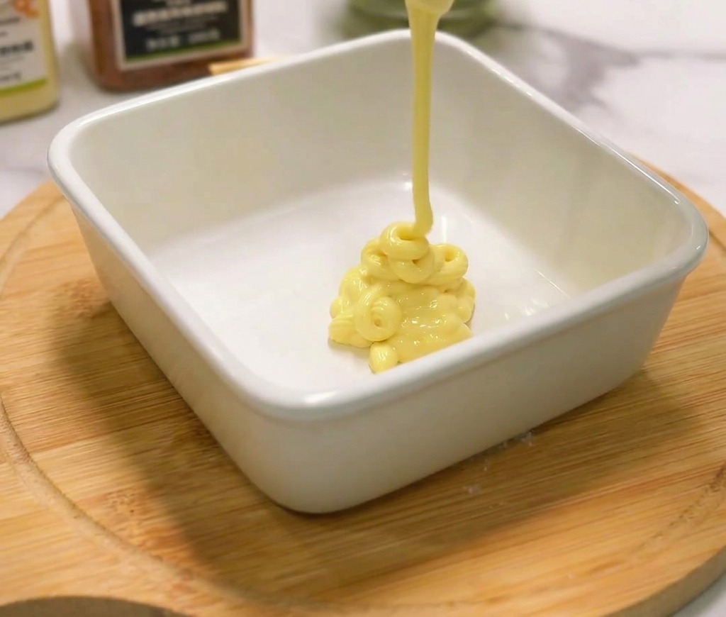 Mayonesa cremosa de color amarillo saliendo de una botella hacia un pequeño tazón de cerámica blanca.