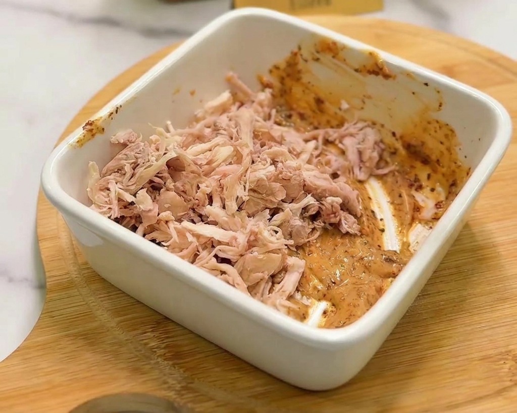 Una montaña de pollo desmechado siendo mezclada con una salsa cremosa naranja en un tazón blanco.
