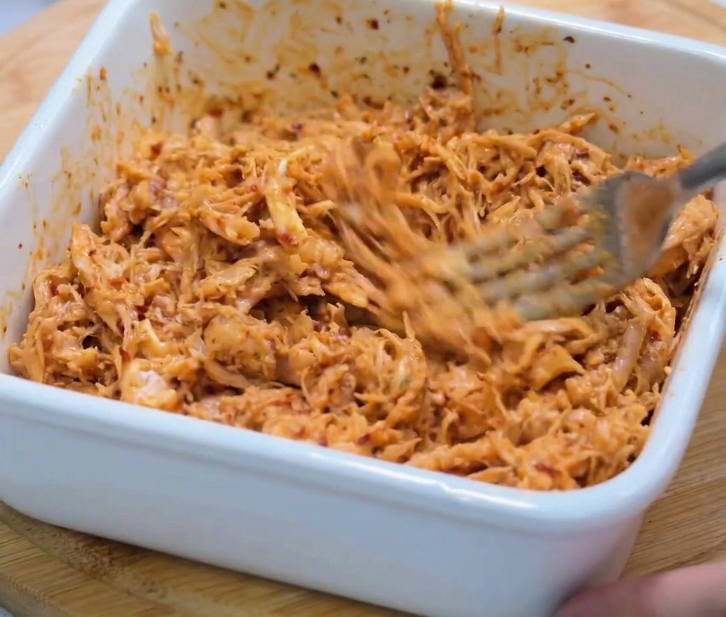 Primer plano de pollo desmechado siendo revuelto en una salsa espesa de color rojo picante con un tenedor de metal.