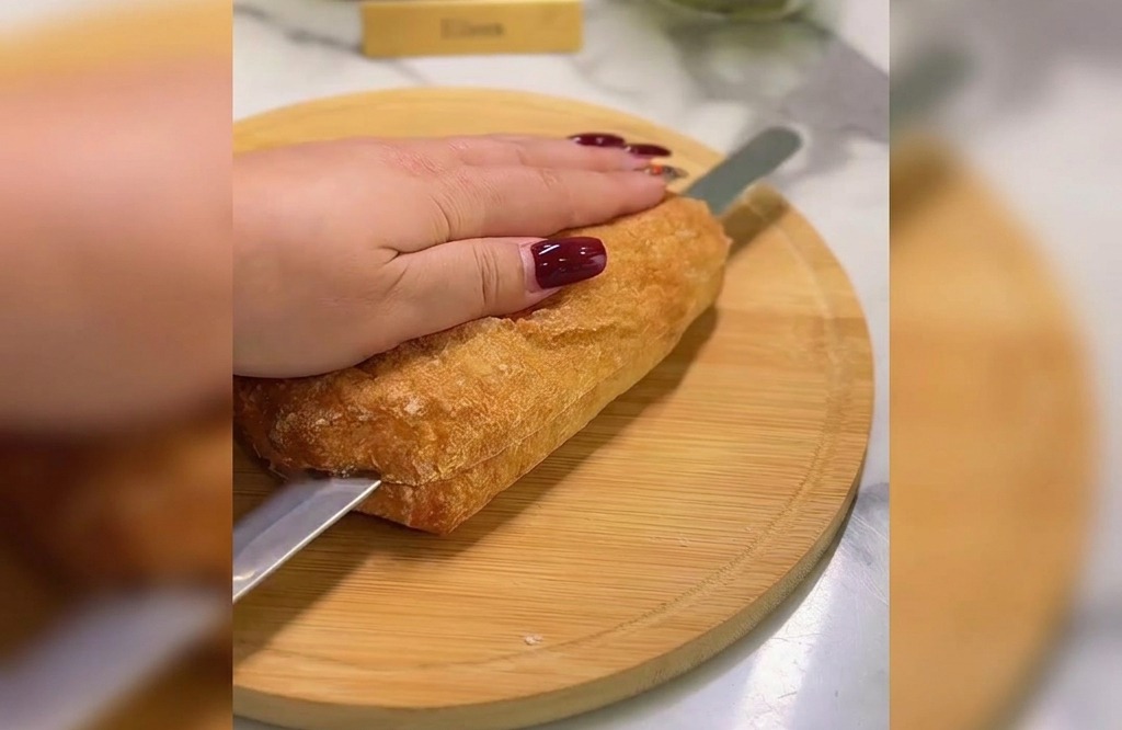Una mano sosteniendo una ciabatta dorada mientras un cuchillo de sierra la corta por la mitad sobre una tabla de madera.
