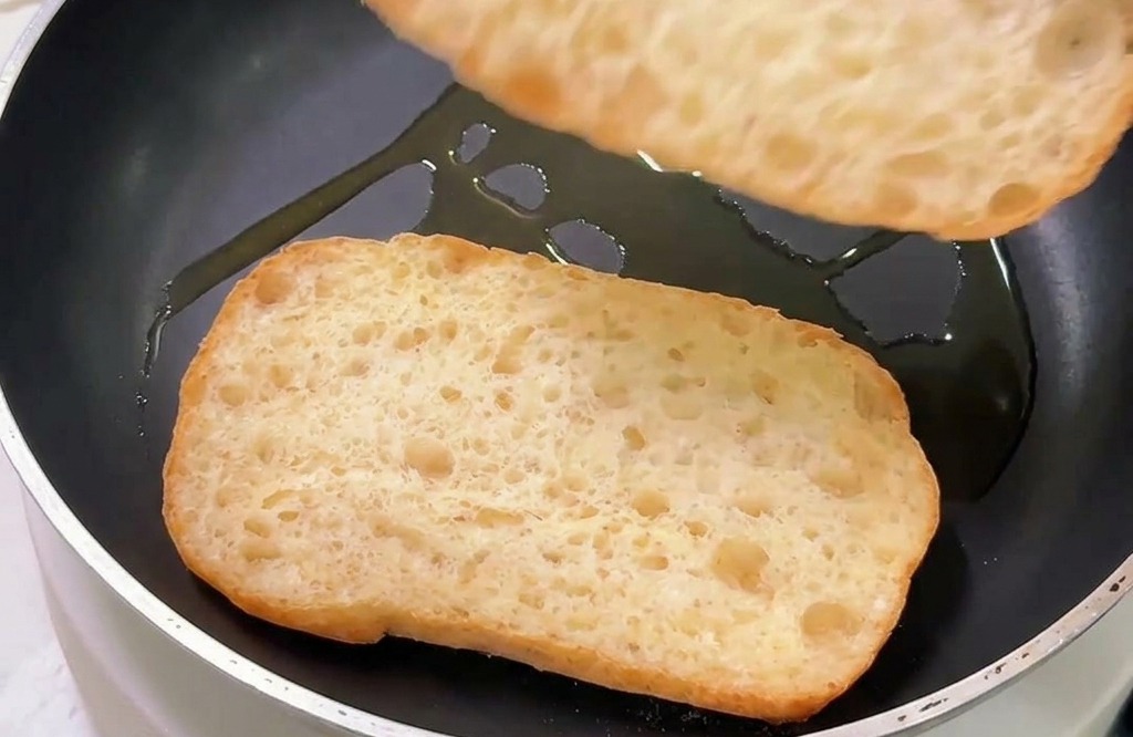 Dos trozos de pan ciabatta tostándose por el lado del corte en una sartén antiadherente con un poco de aceite.