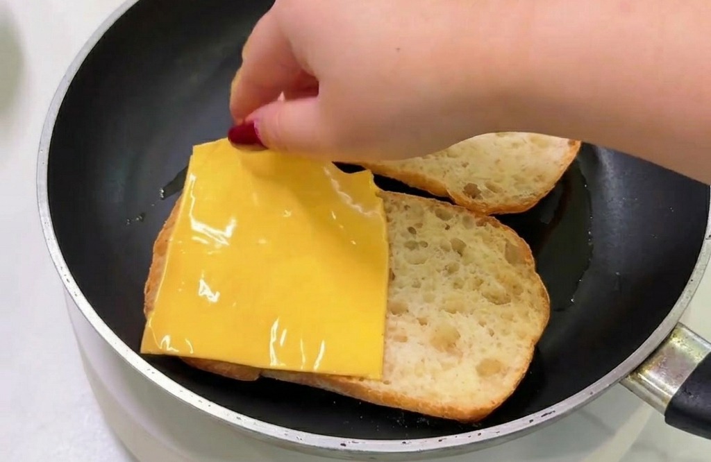Una mano colocando una loncha de queso amarillo sobre un trozo de pan tostado dentro de una sartén.