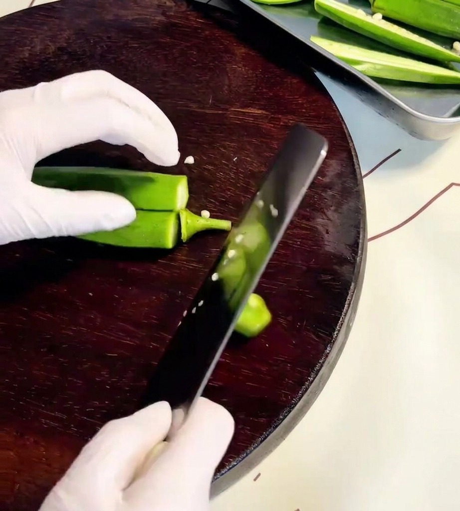Primer plano de manos usando un cuchillo para cortar vainas de okra a lo largo sobre una tabla de cortar de madera.