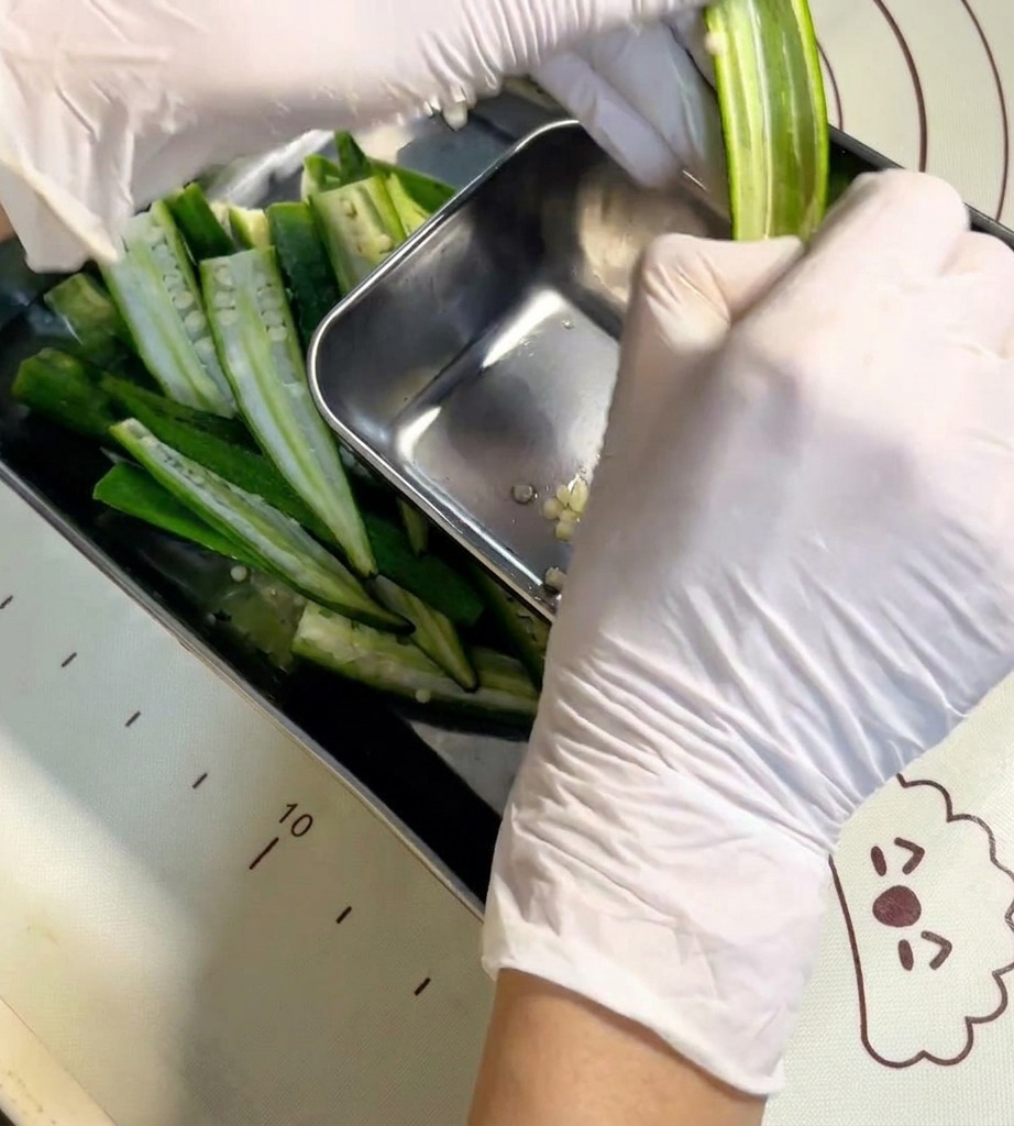 Una mano usando una cuchara pequeña de metal para limpiar las semillas del interior de una vaina de okra abierta.
