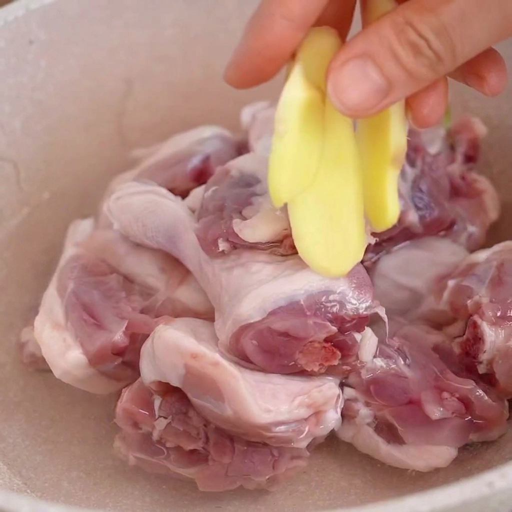 Una mano añadiendo rodajas de jengibre fresco a una olla con trozos de pato crudo sumergidos en agua.
