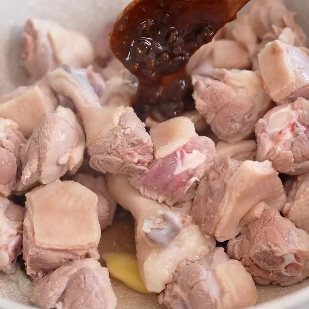 Una cuchara de madera echando pasta oscura de frijoles sobre trozos de pato en una sartén de color claro.