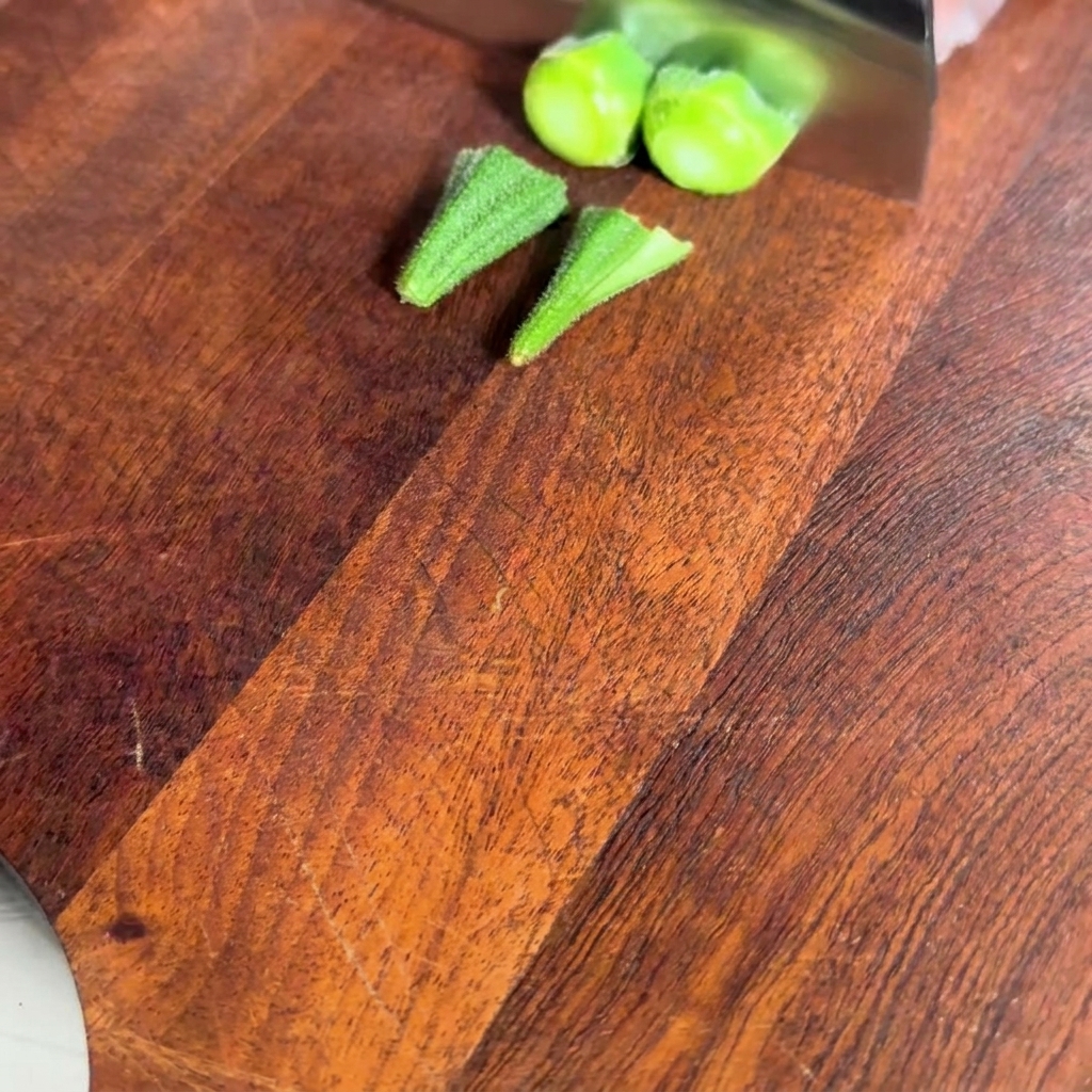 Vista cercana de vainas de okra blanqueadas sobre una tabla de cortar de madera mientras se cortan los extremos del tallo.