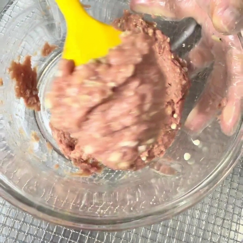 Carne de pato picada y semillas de okra siendo mezcladas en un cuenco con una espátula de silicona amarilla.