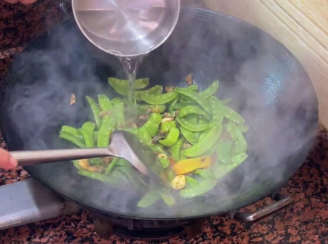 Agua tibia siendo vertida en el wok con los tirabeques y la salchicha salteándose.