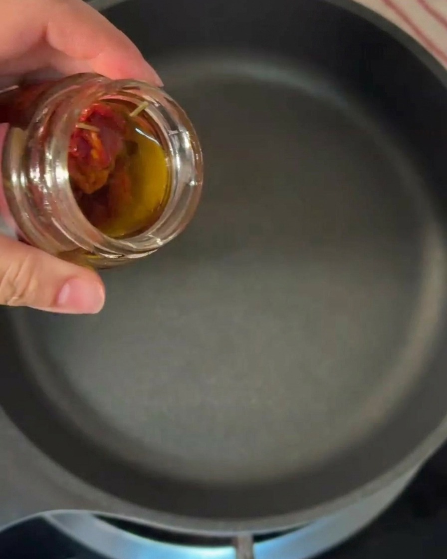 Una mano vertiendo aceite de oliva teñido de naranja desde un pequeño frasco de vidrio en una sartén caliente.
