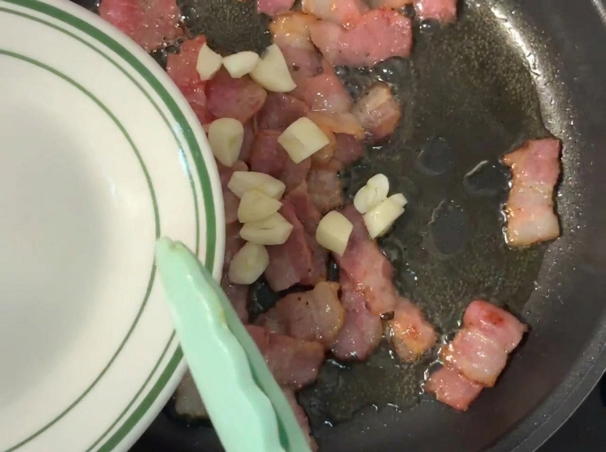 Ajo picado siendo añadido desde un plato a una sartén caliente con trozos de bacon.