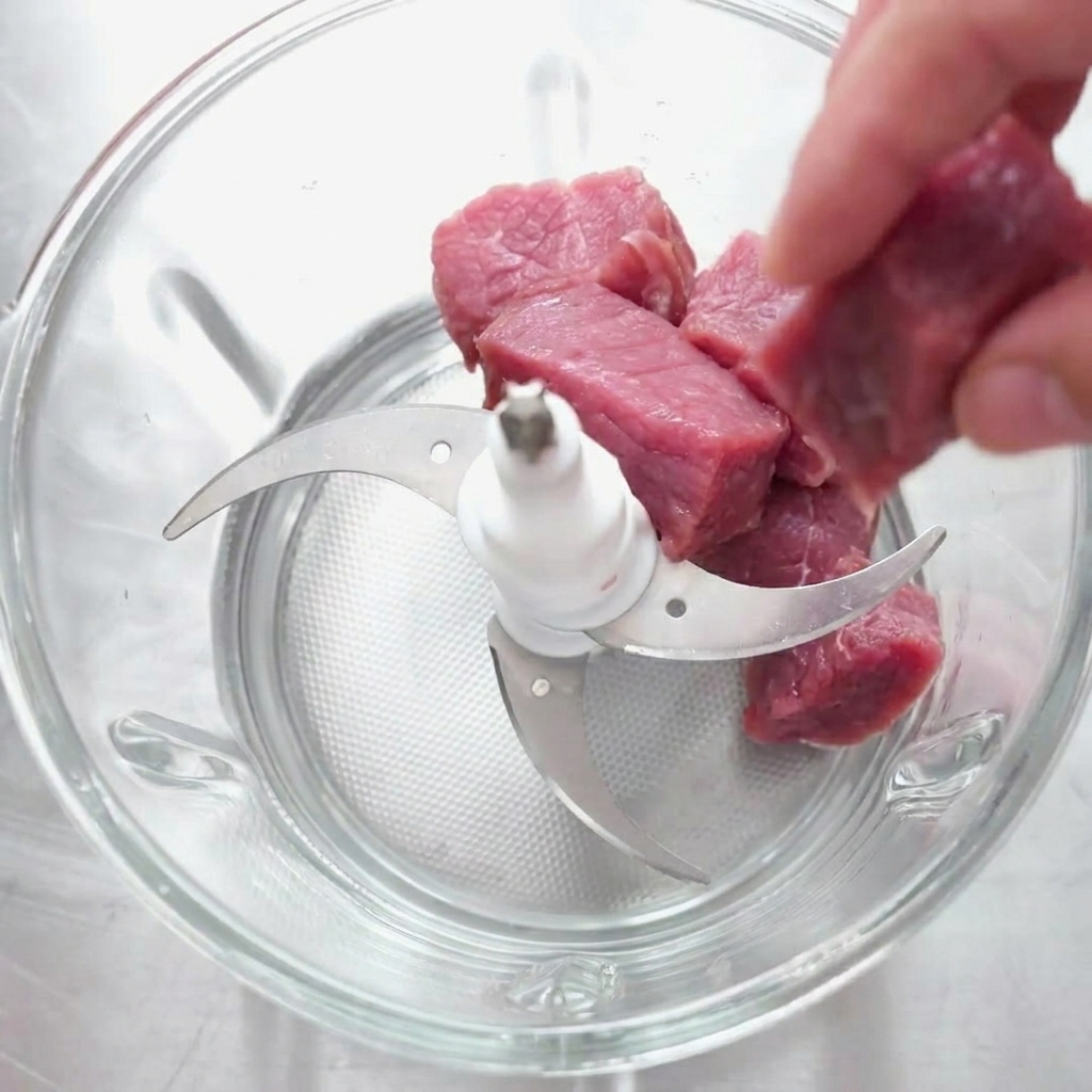 Trozos de carne cruda siendo colocados en el recipiente de vidrio de una picadora de alimentos equipada con una cuchilla múltiple.