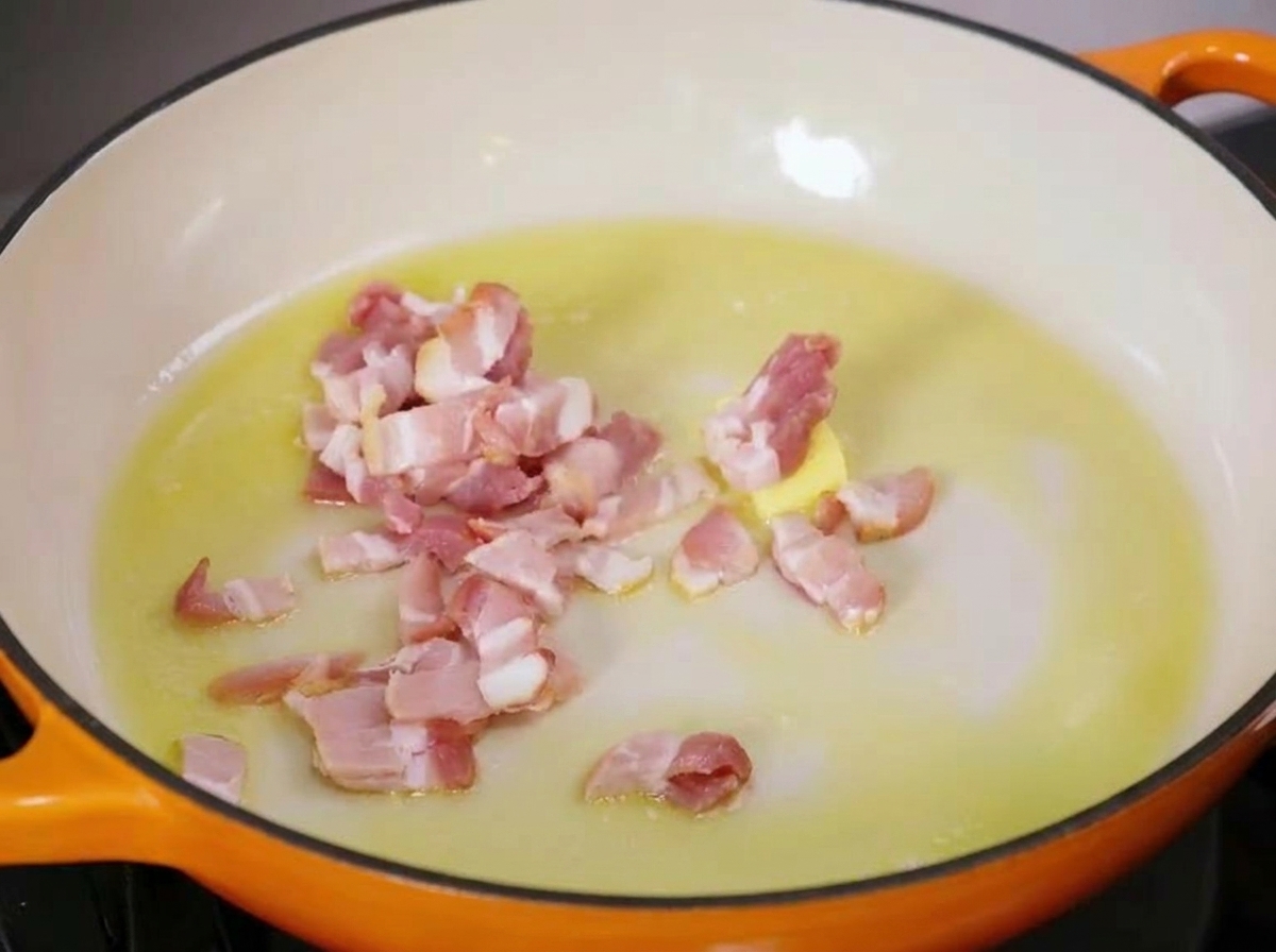 Trozos de tocino crudo chisporroteando en una sartén caliente con mantequilla y aceite de oliva derritiéndose.