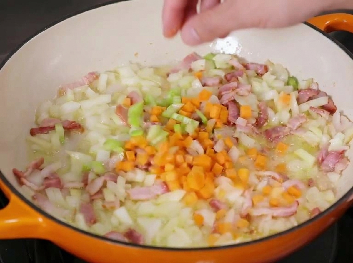 Zanahorias picadas siendo añadidas a una sartén de hierro fundido que contiene cebollas, apio y trozos de tocino ligeramente salteados.