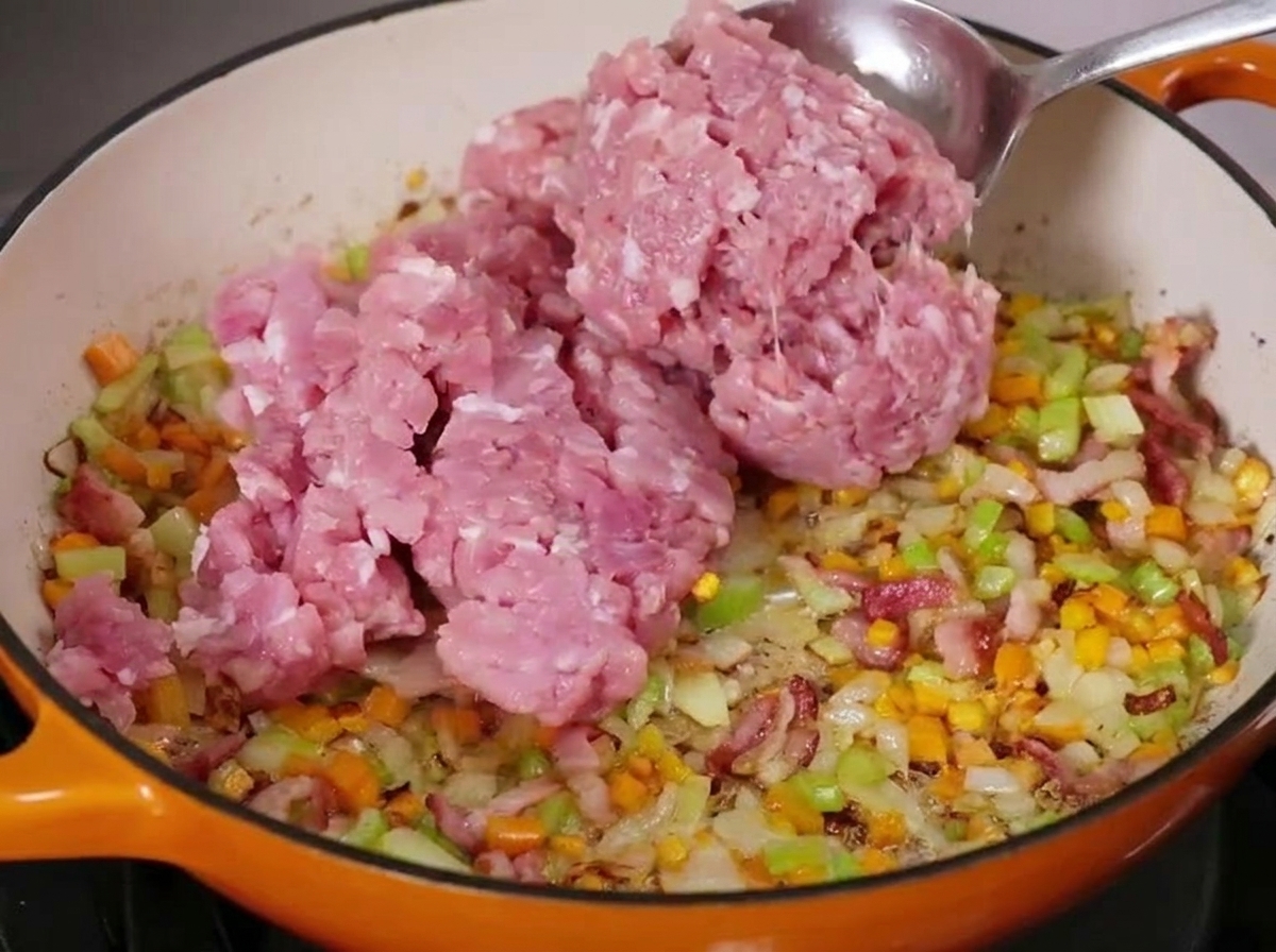 Una gran cucharada de carne picada cruda siendo añadida a una sartén con verduras y tocino.