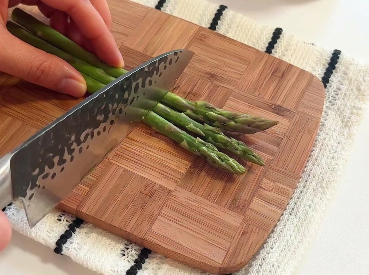 Manos usando un cuchillo de chef para cortar espárragos verdes frescos en trozos cortos sobre una tabla de cortar de madera.
