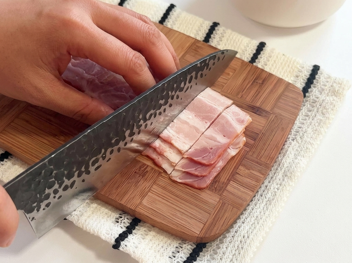 Tiras de bacon crudo siendo cortadas en trozos pequeños sobre una tabla de madera con un cuchillo grande.