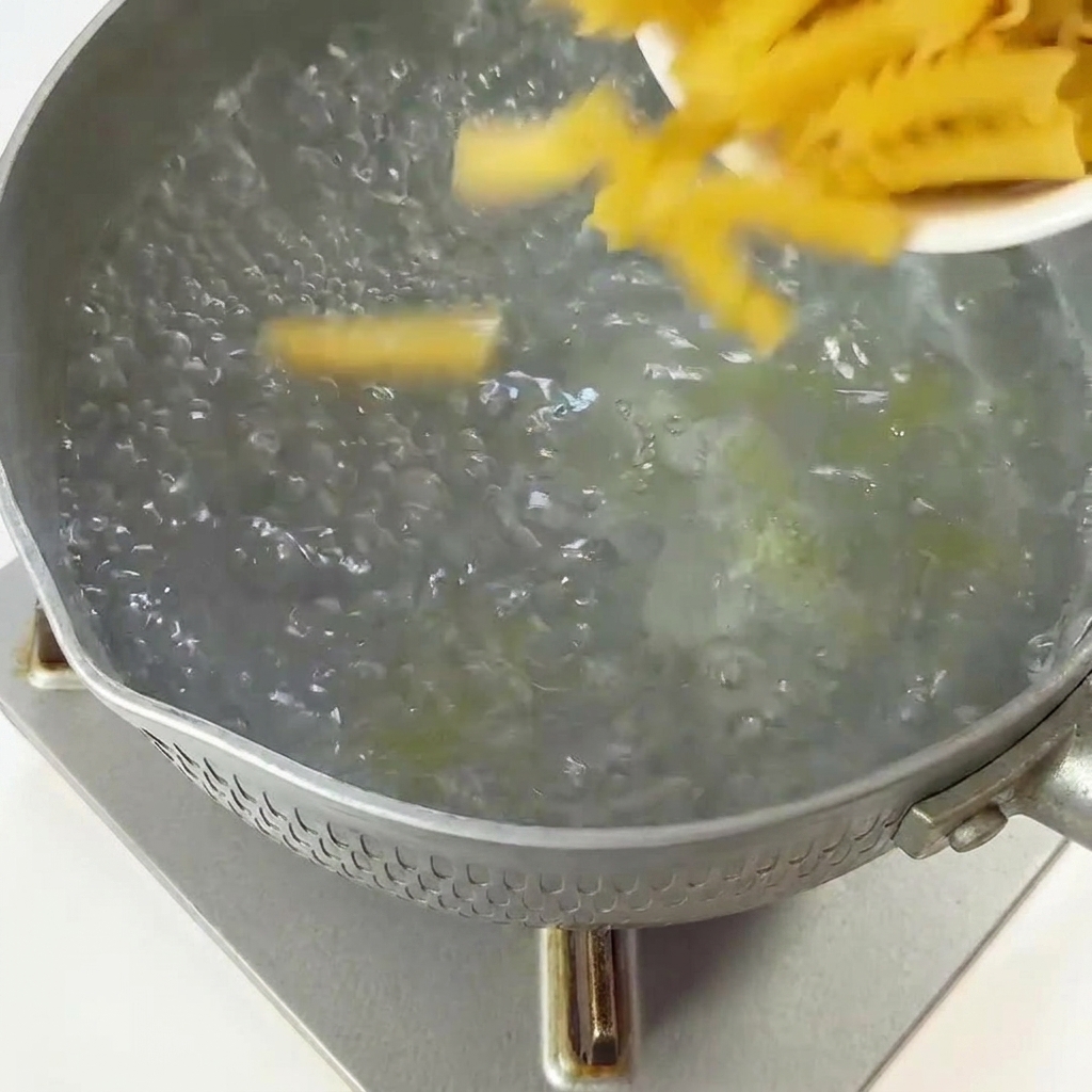 Pasta fusilli seca siendo añadida desde un recipiente pequeño a una olla con agua hirviendo.