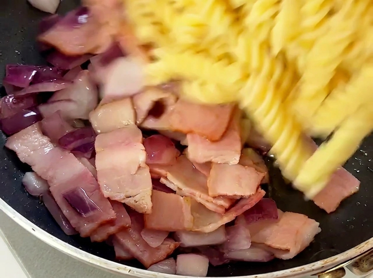 Pasta fusilli cocida siendo añadida a la sartén con el bacon y la cebolla.