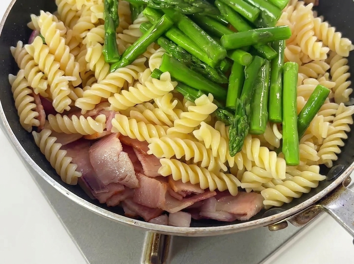 Espárragos y condimentos siendo mezclados con la pasta, el bacon y la cebolla en una sartén.