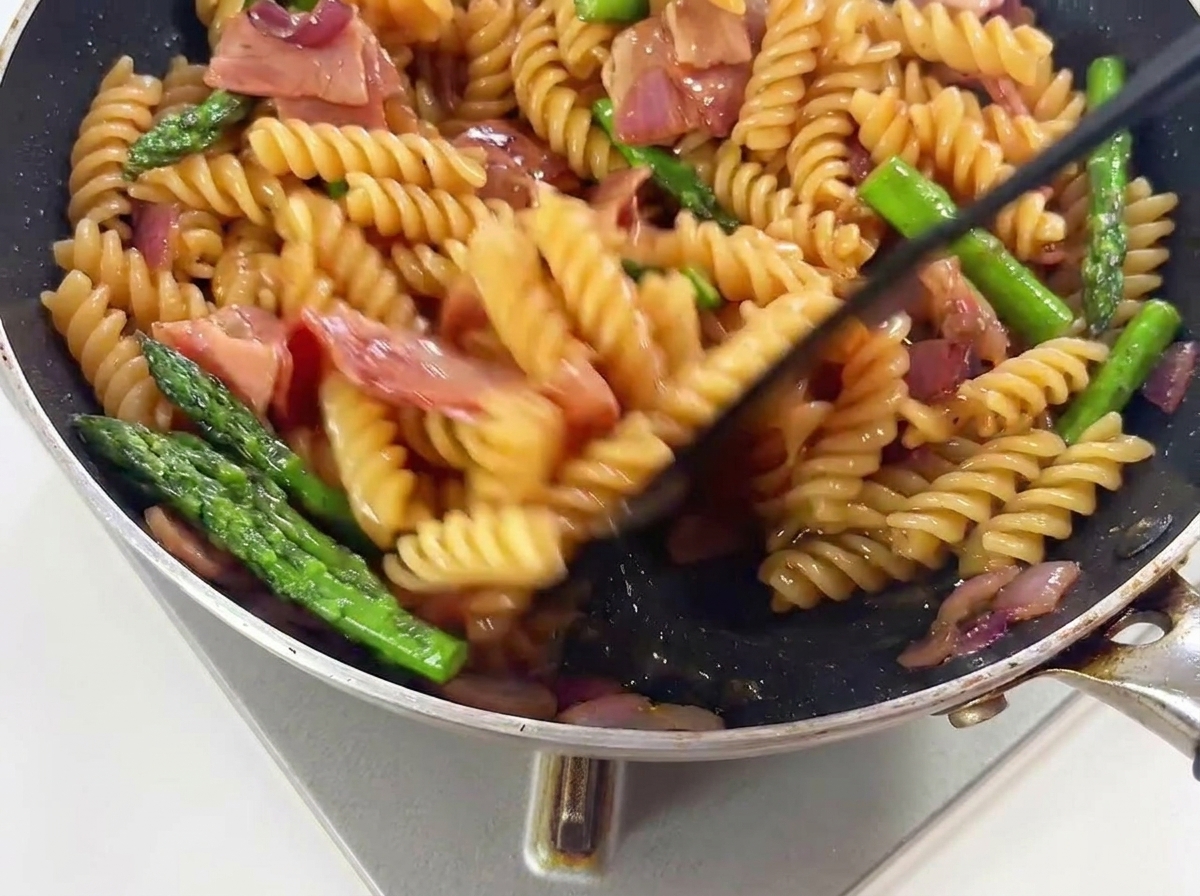 Pasta, espárragos verdes y trozos de bacon siendo mezclados en una sartén caliente para unificar con la salsa.