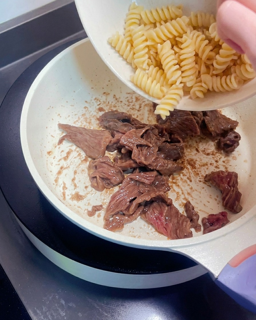 Pasta en espiral siendo transferida desde un bol a una sartén con tiras de ternera cocinadas.