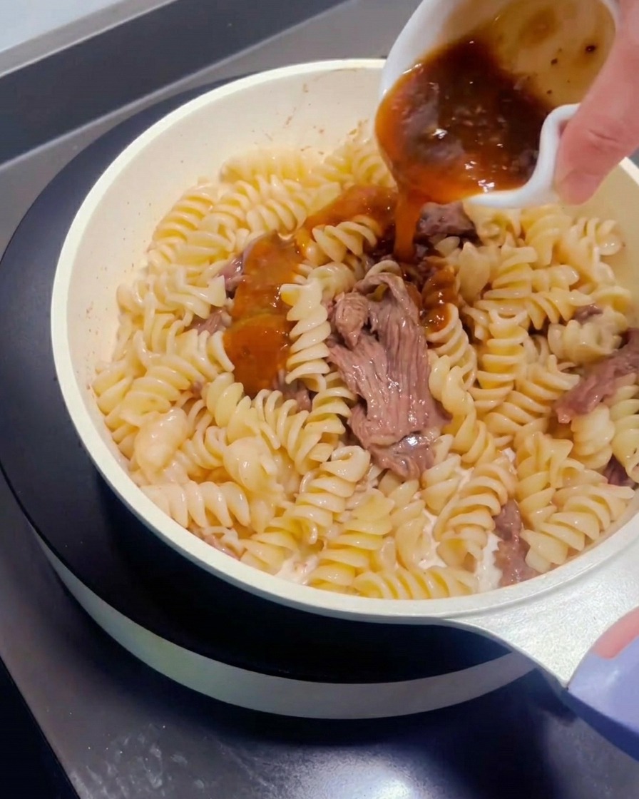 Salsa para pasta siendo vertida desde un pequeño recipiente sobre la ternera y la pasta en la sartén.