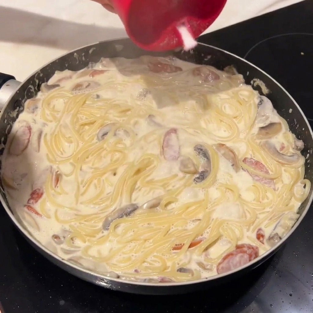 Espaguetis cocidos siendo añadidos a la salsa cremosa en la sartén.