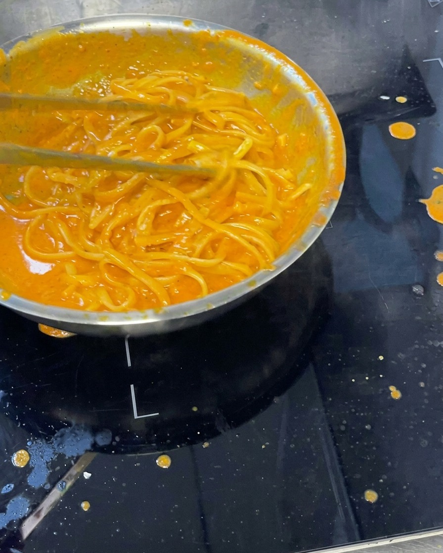 Pasta cremosa mezclada con salsa de pimientos asados en una sartén, con un cucharón mezclando los ingredientes.