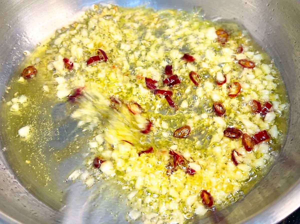 Pasta siendo mezclada en una sartén con ajo dorado y copos de chile, cubriéndose con una salsa brillante y emulsionada.