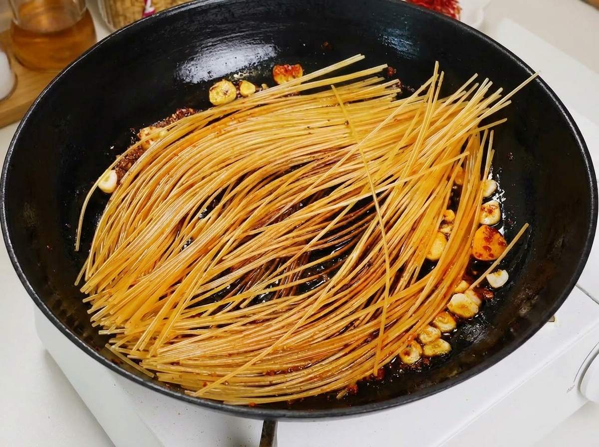 Un cucharón vertiendo caldo de tomate sobre los espaguetis sellados en una sartén negra.
